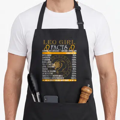 Fact zodiac aprons - Leo Girl Facts nutrition label print with lion icon and orange white text — shown on a black cotton canvas apron against a white background