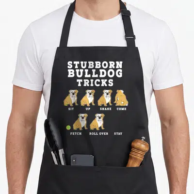 Trick dog apron - Tan and white bulldogs ignoring sit stay fetch commands under Stubborn Bulldog Tricks text — shown on a black cotton canvas apron against a white background