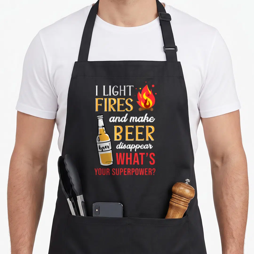 Light camping apron - I light fires and make beer disappear text with campfire and bottle illustration — shown on a black cotton canvas apron against a white background