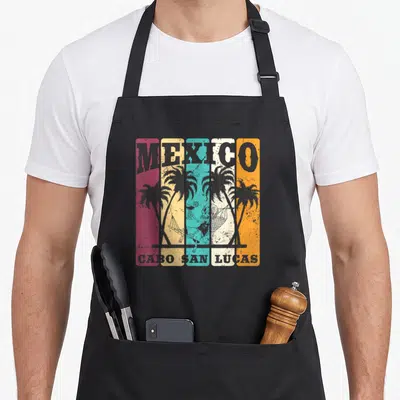 San lucas souvenir mexico apron - Retro skeleton in hammock with palm trees across five colored stripes and bold MEXICO — shown on a black cotton canvas apron against a white background