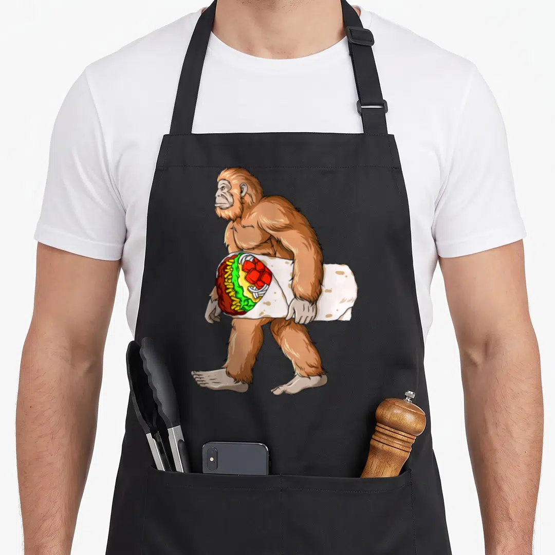 Bigfoot mexican apron - Brown cartoon sasquatch walking left carrying a large burrito with visible red green and yellow — shown on a black cotton canvas apron against a white background