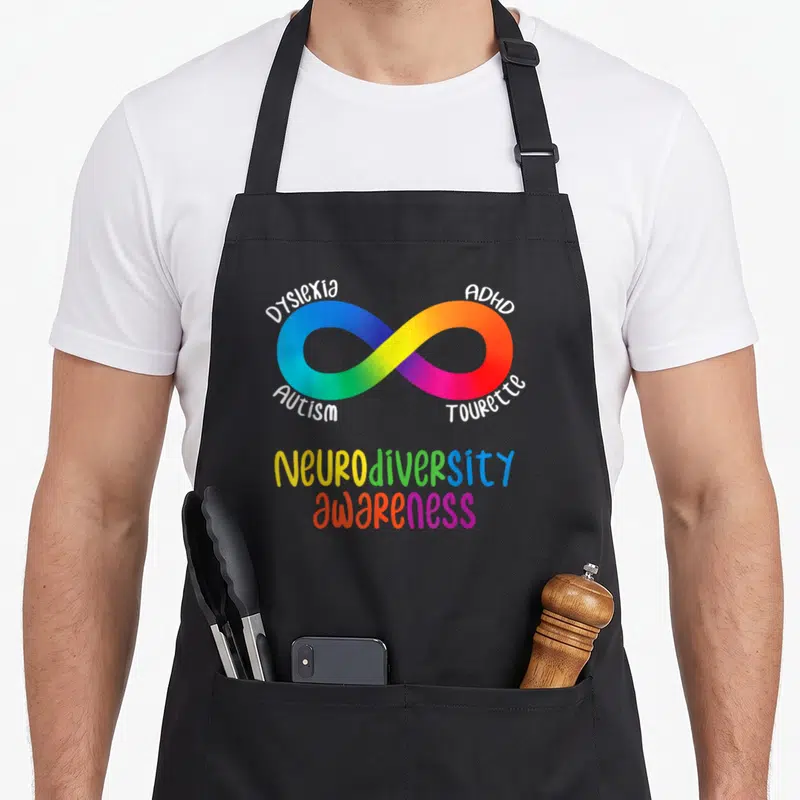 Neurodiversity tourette awareness apron - Rainbow infinity symbol surrounded by Dyslexia ADHD Autism Tourette text — shown on a black cotton canvas apron against a white background