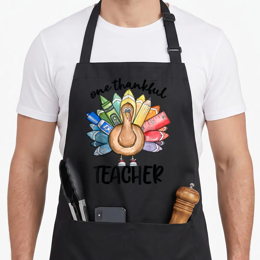 Teacher thankful apron - Cartoon turkey with crayon pencil ruler tail feathers and one thankful TEACHER text — shown on a black cotton canvas apron against a white background