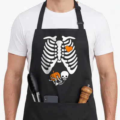 Pregnancy halloween apron - White skeleton ribcage with orange heart above pumpkin skull and bones cartoon — shown on a black cotton canvas apron against a white background