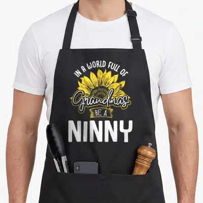 Ninny grandma's aprons - Yellow sunflower with stacked white text IN A WORLD FULL OF Grandmas BE A NINNY — shown on a black cotton canvas apron against a white background