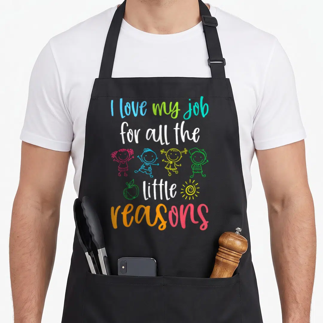 School reason apron - Stacked colorful text I love my job with four cartoon children apple and sun — shown on a black cotton canvas apron against a white background