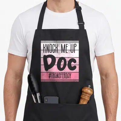 Knock infertility awareness apron - Grey KNOCK ME UP DOC text on pink and white horizontal stripes — shown on a black cotton canvas apron against a white background