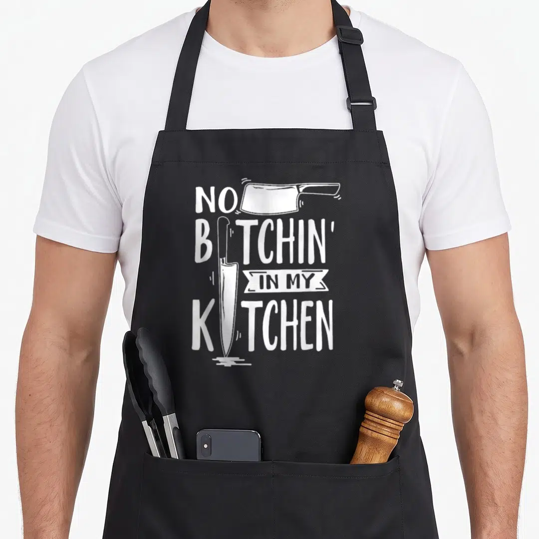 Bitchin executive chef apron - White cleaver and chef knife integrated into NO BITCHIN IN MY KITCHEN typography — shown on a black cotton canvas apron against a white background