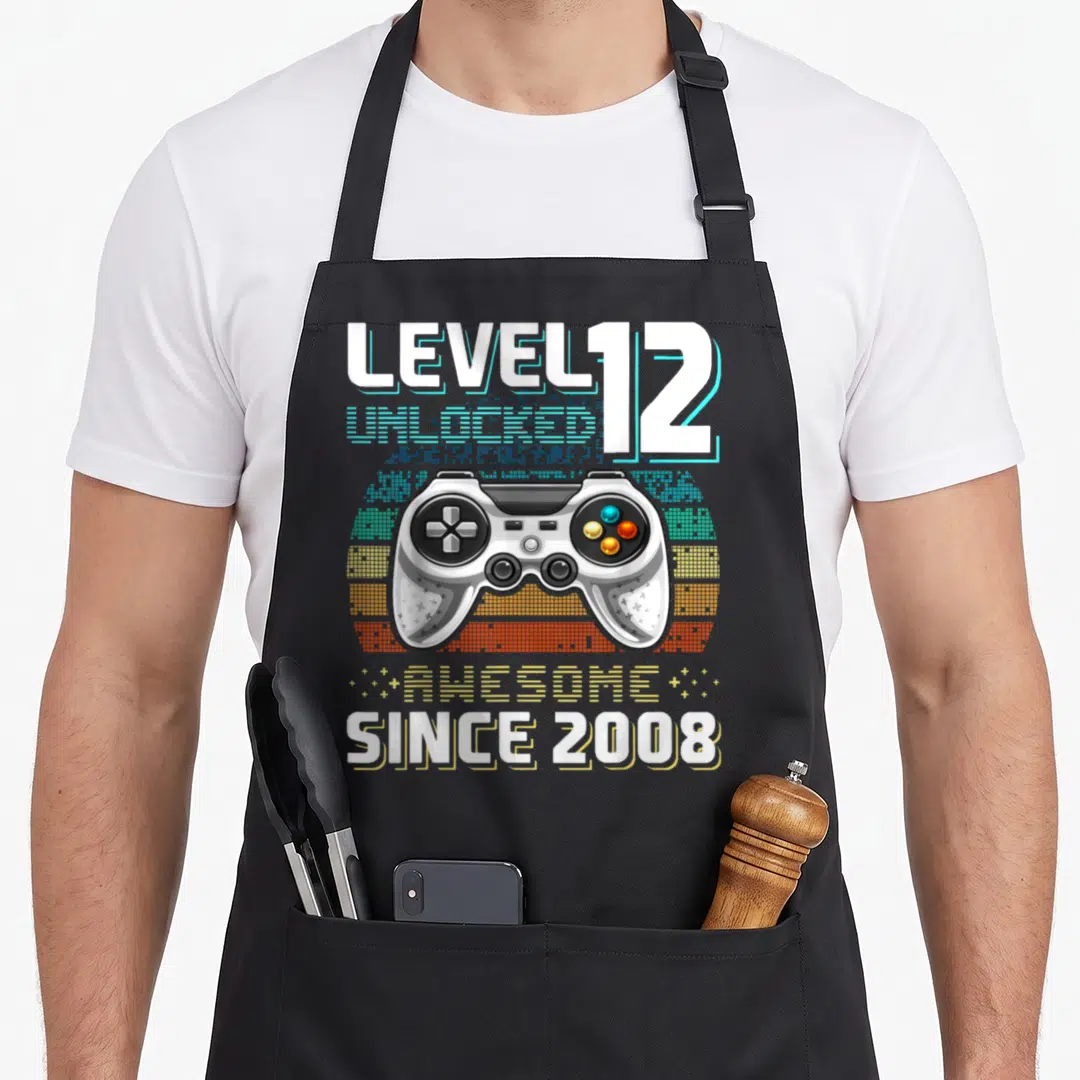 12th video game apron - Retro game controller with pixelated sunburst and text Level 12 Unlocked Awesome Since 2008 — shown on a black cotton canvas apron against a white background