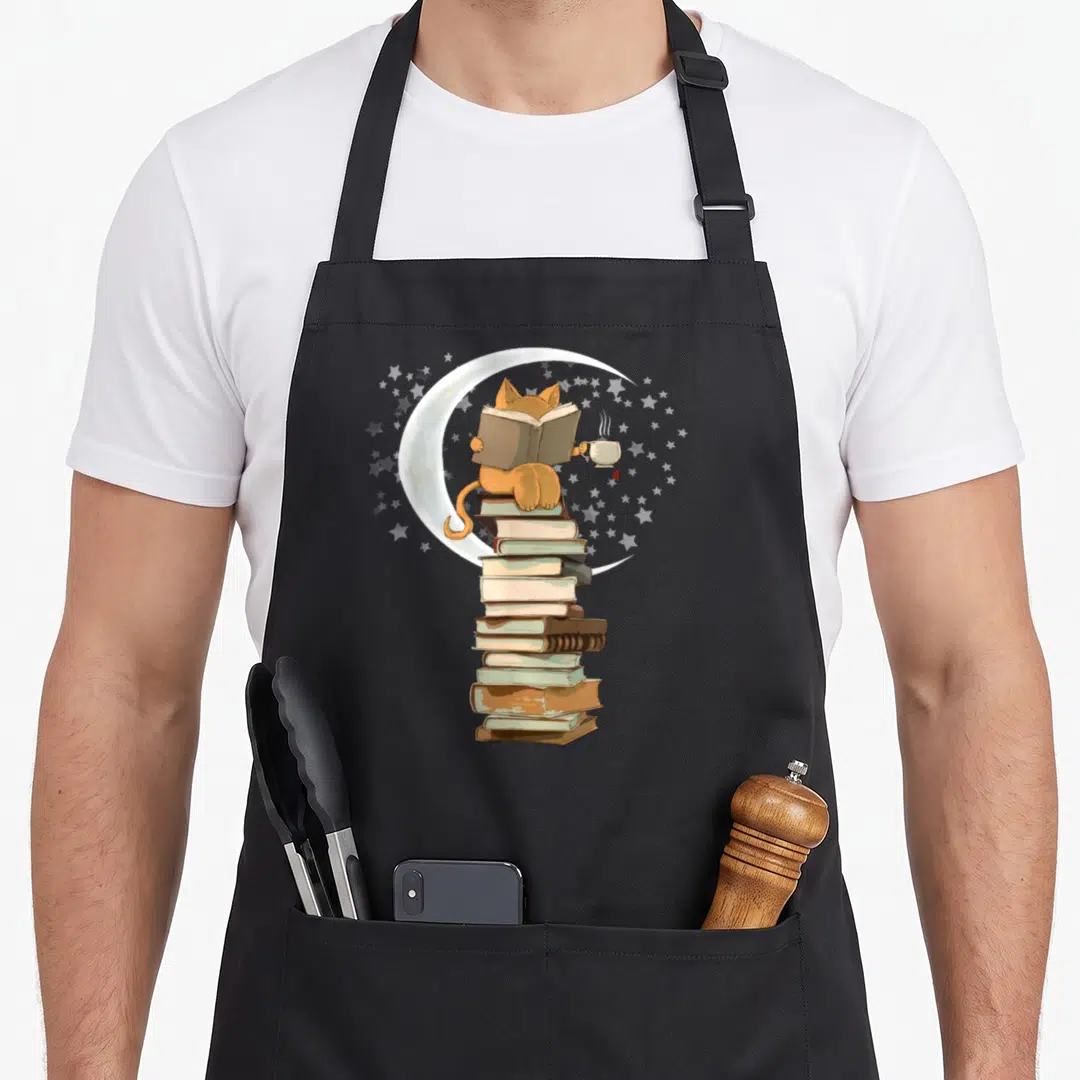 Moonlight cat apron - Orange cat sitting on stacked books reading under a crescent moon with stars — shown on a black cotton canvas apron against a white background