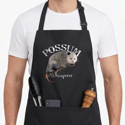 Whisperer possum apron - Photo-style opossum on branch with arched POSSUM and script Whisperer text — shown on a black cotton canvas apron against a white background
