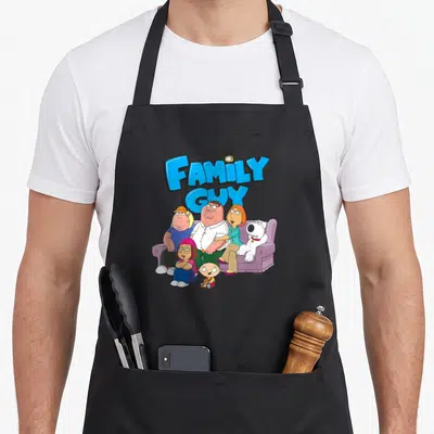 Griffin cartoon apron - Family Guy characters on purple sofa below stacked blue text with small TV on top of word FAMILY — shown on a black cotton canvas apron against a white background