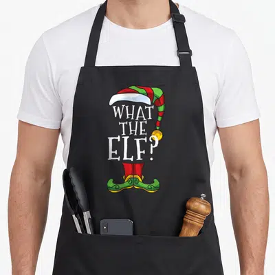 Elve elf apron - Cartoon elf hat and shoes framing white WHAT THE ELF? text in red green and yellow — shown on a black cotton canvas apron against a white background