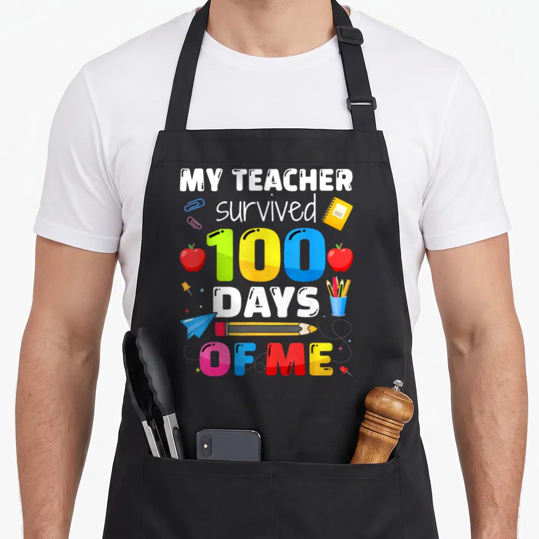 Survived 100th teacher apron - My teacher survived 100 days of me text with apples and stationery — shown on a black cotton canvas apron against a white background