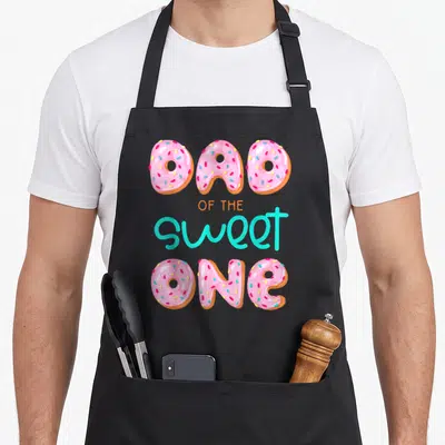 Sweet family aprons - Donuts spelling DAD OF THE sweet ONE in pink turquoise orange and brown cartoon style — shown on a black cotton canvas apron against a white background