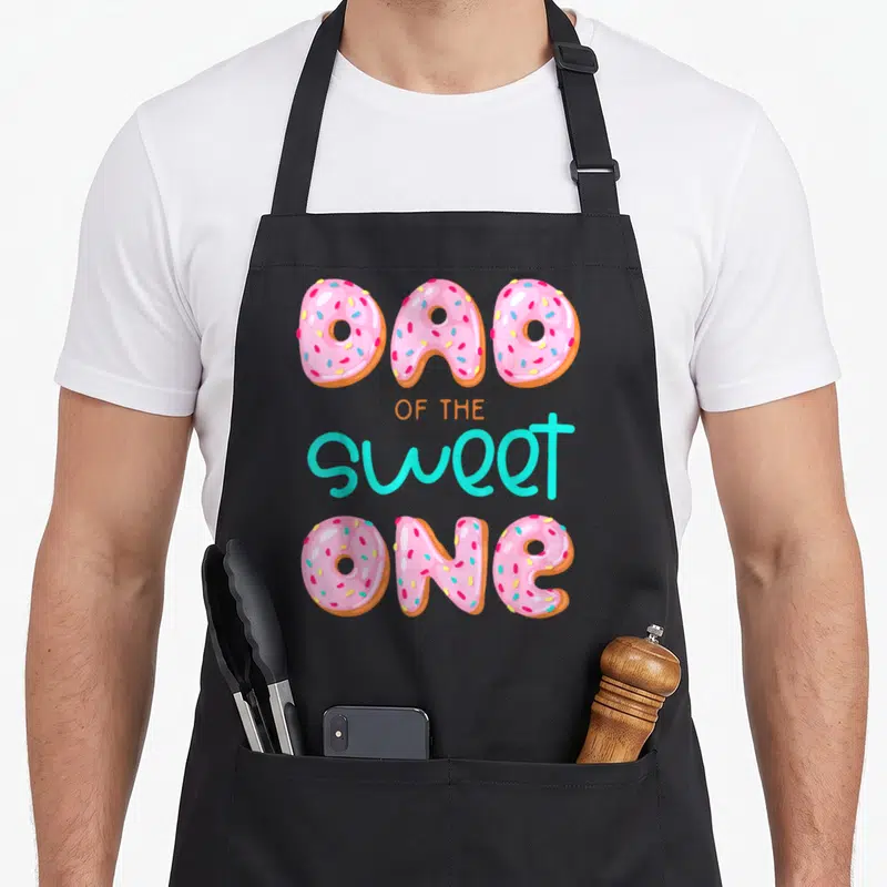 Sweet family aprons - Donuts spelling DAD OF THE sweet ONE in pink turquoise orange and brown cartoon style — shown on a black cotton canvas apron against a white background