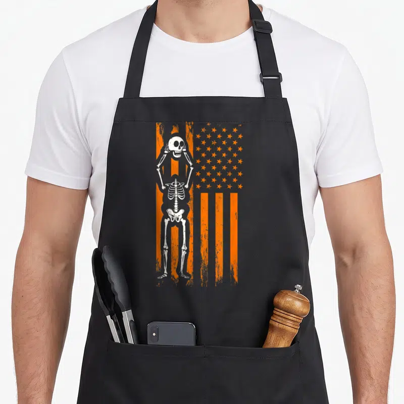 Spooky american flag apron - White skeleton standing before distressed orange and grey striped flag with stars — shown on a black cotton canvas apron against a white background