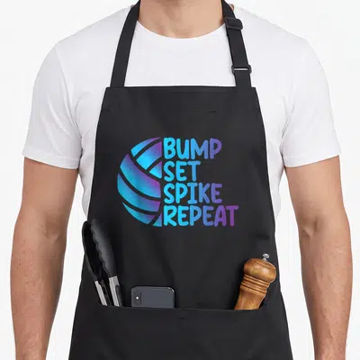 Volleyball bump apron - Stylized ball in blue and purple next to stacked text BUMP SET SPIKE REPEAT — shown on a black cotton canvas apron against a white background