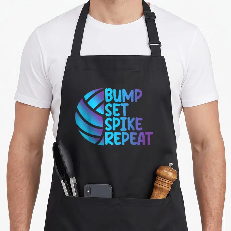 Volleyball bump apron - Stylized ball in blue and purple next to stacked text BUMP SET SPIKE REPEAT — shown on a black cotton canvas apron against a white background