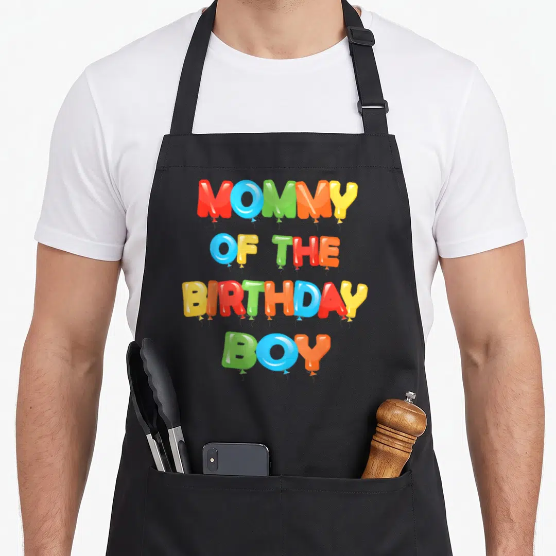 Funny birthday balloon apron - MOMMY OF THE BIRTHDAY BOY text formed by red blue green orange and yellow cartoon — shown on a black cotton canvas apron against a white background