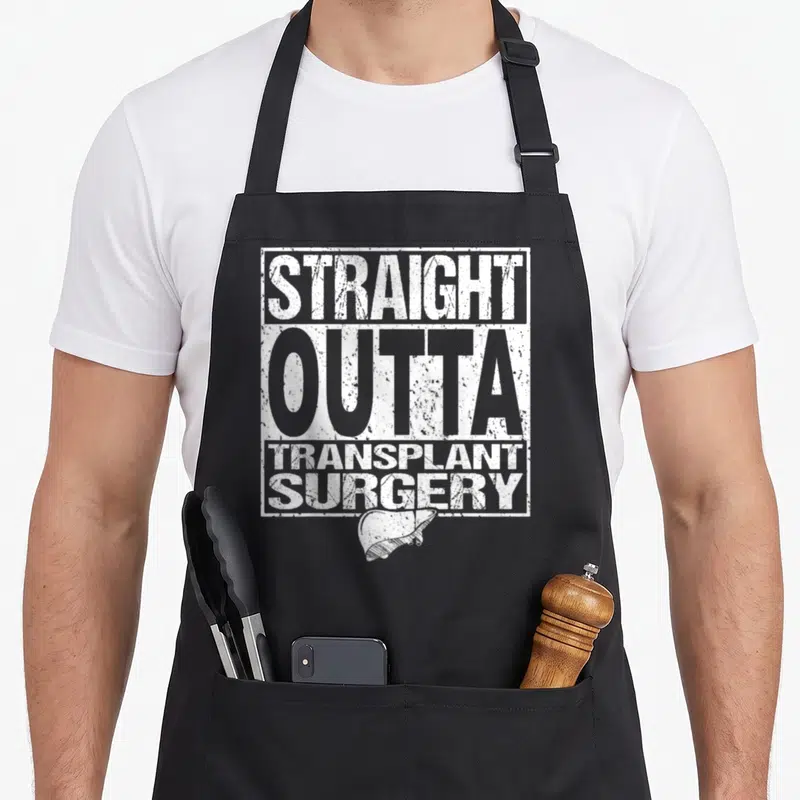 Outta organ apron - Distressed white text Straight Outta Transplant Surgery above a liver drawing on black bib — shown on a black cotton canvas apron against a white background