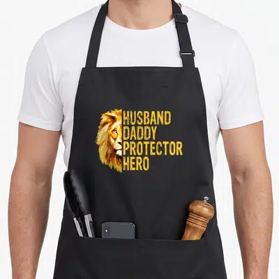 Lion daddy aprons - Half lion face beside stacked yellow text HUSBAND DADDY PROTECTOR HERO — shown on a black cotton canvas apron against a white background