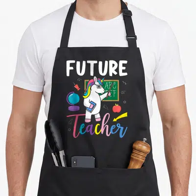 Future school teacher apron - White unicorn with glasses holding pointer and book near chalkboard with ABC DEF — shown on a black cotton canvas apron against a white background