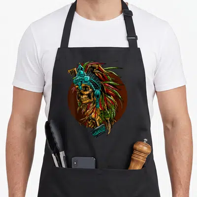 Aztec skeleton apron - Cartoon skull with ornate teal headdress and feathers on brown circle background — shown on a black cotton canvas apron against a white background
