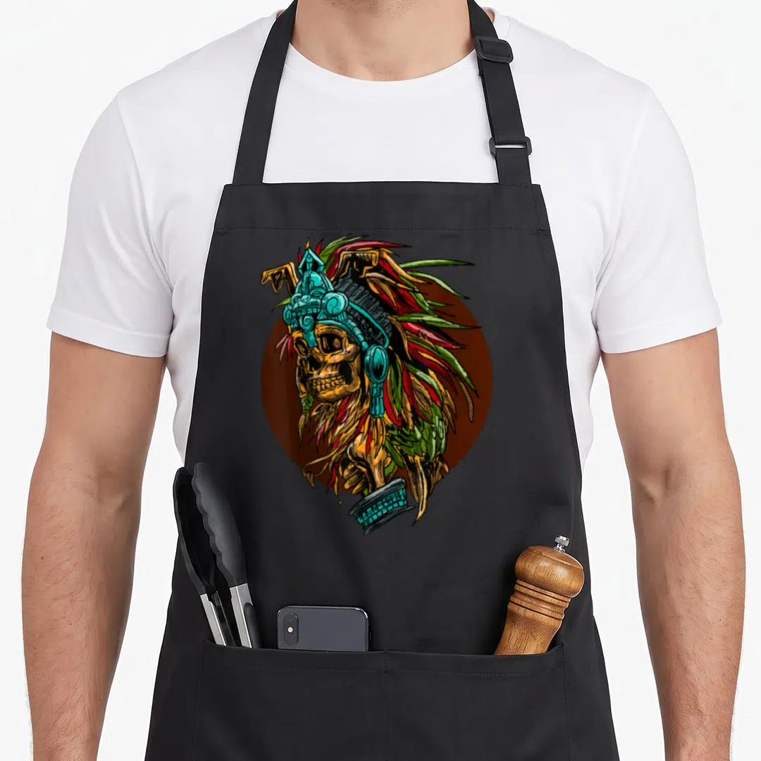 Aztec skeleton apron - Cartoon skull with ornate teal headdress and feathers on brown circle background — shown on a black cotton canvas apron against a white background