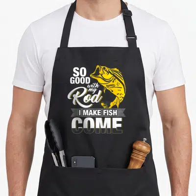 Rod fish apron - Yellow cartoon fish graphic with white and black typography reading So Good With My Rod I Make Fish — shown on a black cotton canvas apron against a white background