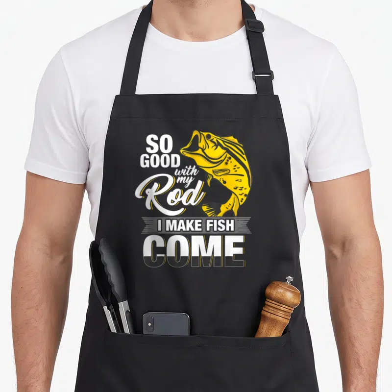 Rod fish apron - Yellow cartoon fish graphic with white and black typography reading So Good With My Rod I Make Fish — shown on a black cotton canvas apron against a white background