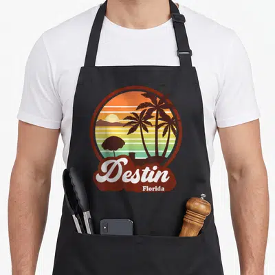 Destin florida tree apron - Circular retro sunset with palm trees, umbrella, and white Destin Florida script on brown — shown on a black cotton canvas apron against a white background