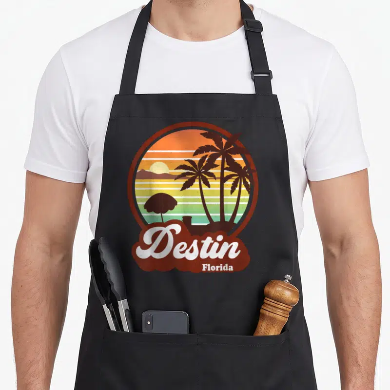 Destin florida tree apron - Circular retro sunset with palm trees, umbrella, and white Destin Florida script on brown — shown on a black cotton canvas apron against a white background