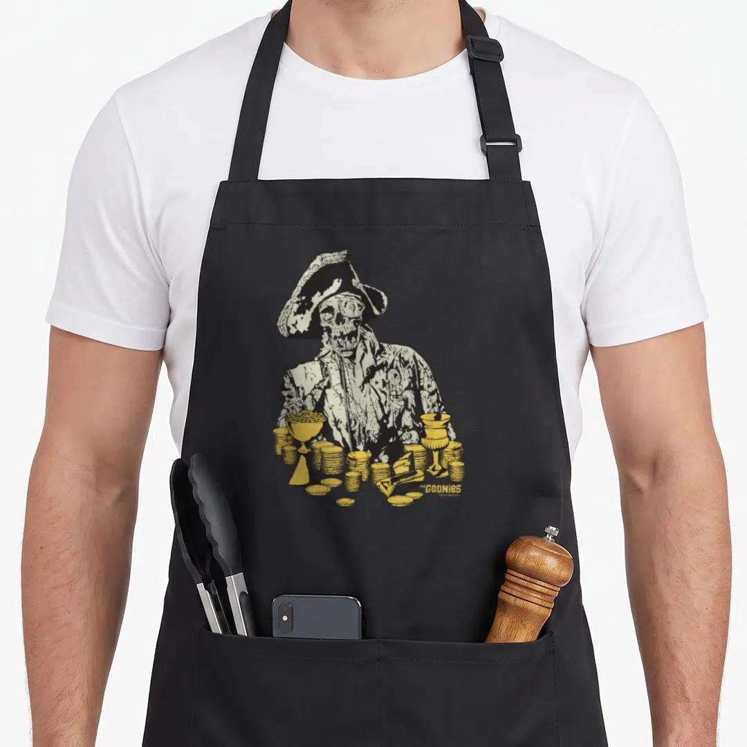 Eyed crazy robin man apron - Skeleton pirate in line art holding yellow gold coins and goblets with The GOONIES logo — shown on a black cotton canvas apron against a white background