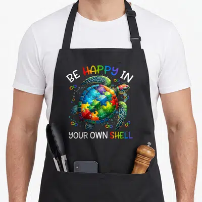 Puzzle turtle dad apron - Rainbow puzzle piece turtle with Be Happy In Your Own Shell text and infinity symbols — shown on a black cotton canvas apron against a white background