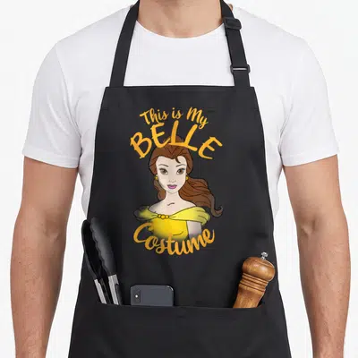 Belle halloween apron - Cartoon Belle in yellow dress with This is My BELLE Costume text in orange and brown — shown on a black cotton canvas apron against a white background