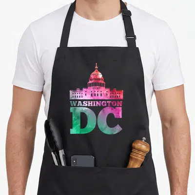 Capital capitol hill apron - Watercolor US Capitol building with stacked WASHINGTON DC text in pink, red, green — shown on a black cotton canvas apron against a white background