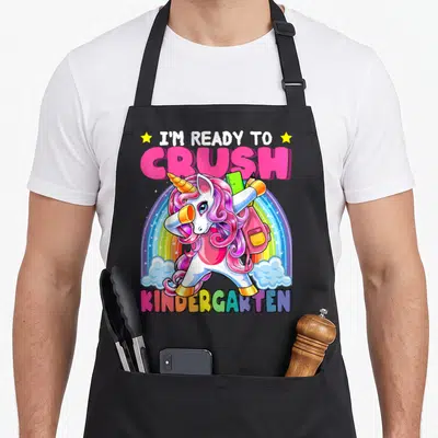 Crush unicorn apron - Dabbing unicorn with backpack and ruler under rainbow text I'm ready to crush kindergarten — shown on a black cotton canvas apron against a white background