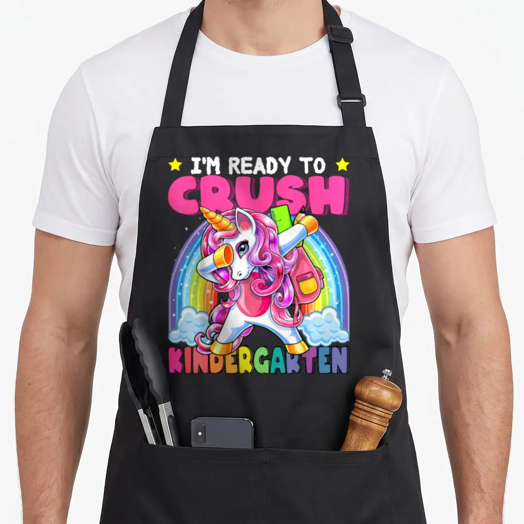Crush unicorn apron - Dabbing unicorn with backpack and ruler under rainbow text I'm ready to crush kindergarten — shown on a black cotton canvas apron against a white background