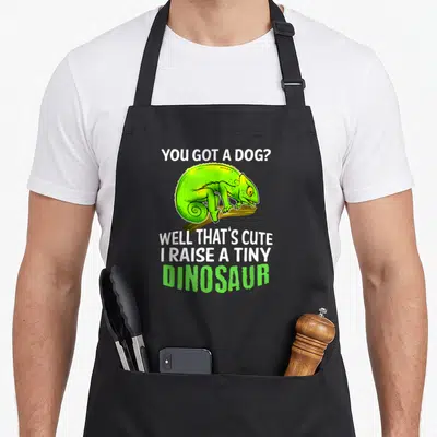Chameleon pet apron - Green cartoon chameleon on branch with white and green dinosaur slogan text — shown on a black cotton canvas apron against a white background