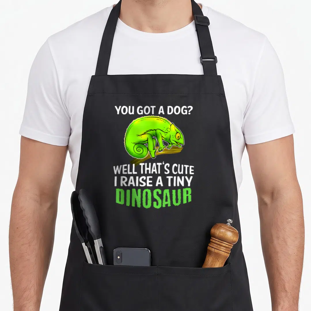 Chameleon pet apron - Green cartoon chameleon on branch with white and green dinosaur slogan text — shown on a black cotton canvas apron against a white background