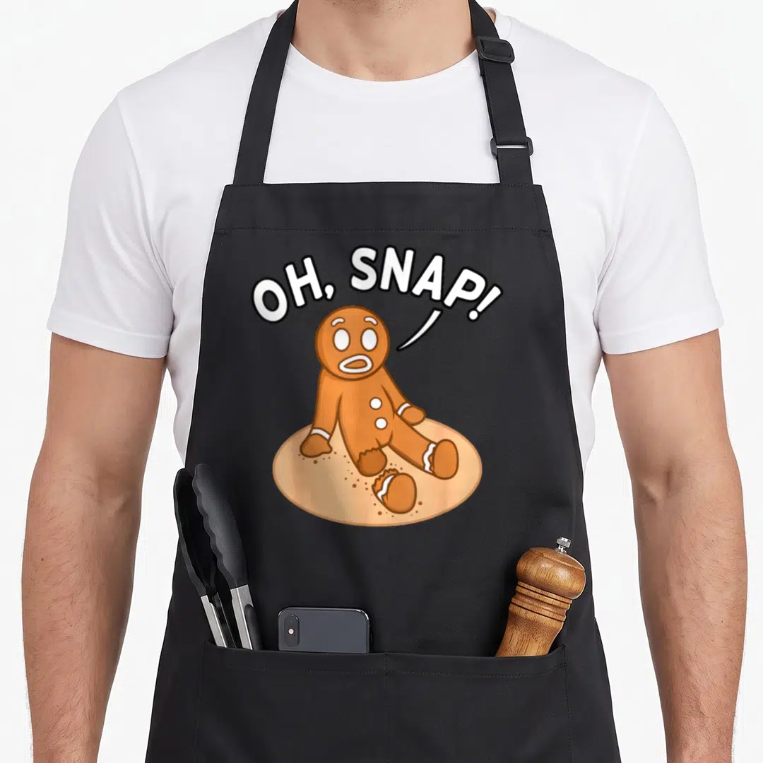 Freak snap aprons - Cartoon gingerbread man with broken leg and OH SNAP speech bubble in brown and tan — shown on a black cotton canvas apron against a white background
