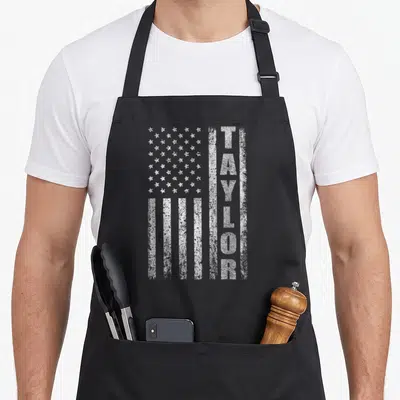 Taylor american flag apron - Vertical white TAYLOR text beside distressed white USA flag stars and stripes — shown on a black cotton canvas apron against a white background