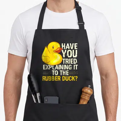 Programmer rubber apron - Yellow cartoon rubber duck next to stacked white text with circuit board lines — shown on a black cotton canvas apron against a white background