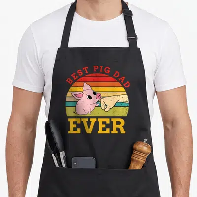 Pig domestic aprons - Cartoon pig and human hand fist bumping under arched BEST PIG DAD text over striped sunset — shown on a black cotton canvas apron against a white background