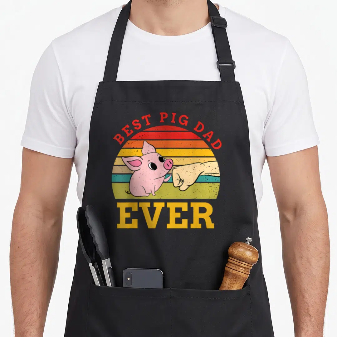 Pig domestic aprons - Cartoon pig and human hand fist bumping under arched BEST PIG DAD text over striped sunset — shown on a black cotton canvas apron against a white background