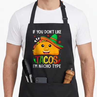 Taco meme apron - Cartoon taco in sombrero with IF YOU DON'T LIKE, TACOS, I'M NACHO TYPE text and chili peppers — shown on a black cotton canvas apron against a white background