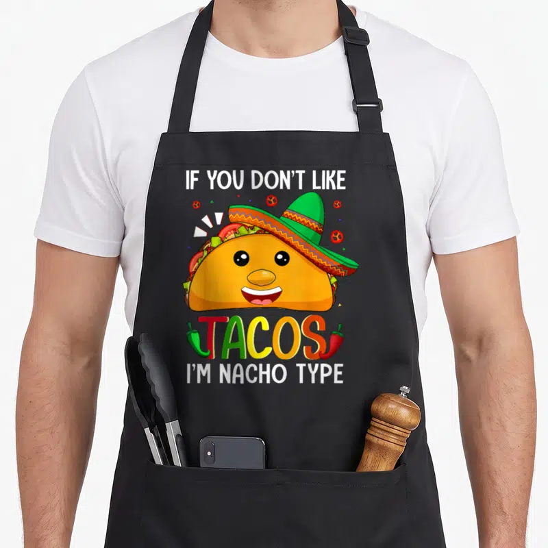 Taco meme apron - Cartoon taco in sombrero with IF YOU DON'T LIKE, TACOS, I'M NACHO TYPE text and chili peppers — shown on a black cotton canvas apron against a white background