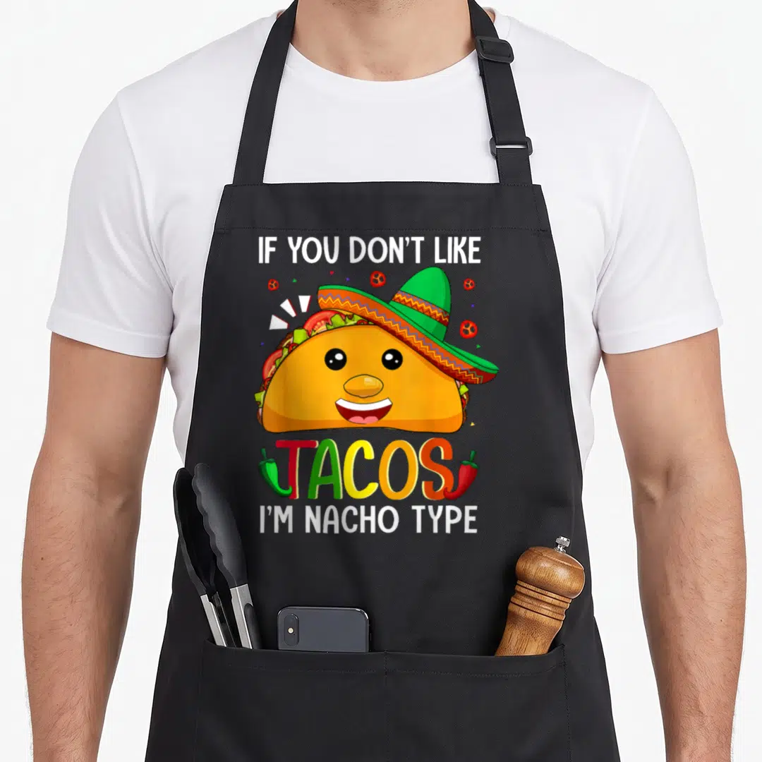 Taco meme apron - Cartoon taco in sombrero with IF YOU DON'T LIKE, TACOS, I'M NACHO TYPE text and chili peppers — shown on a black cotton canvas apron against a white background