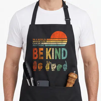 Sign choose kindness be kind inspirational apron - Retro sun and six colorful sign language hands below stacked 'BE — shown on a black cotton canvas apron against a white background